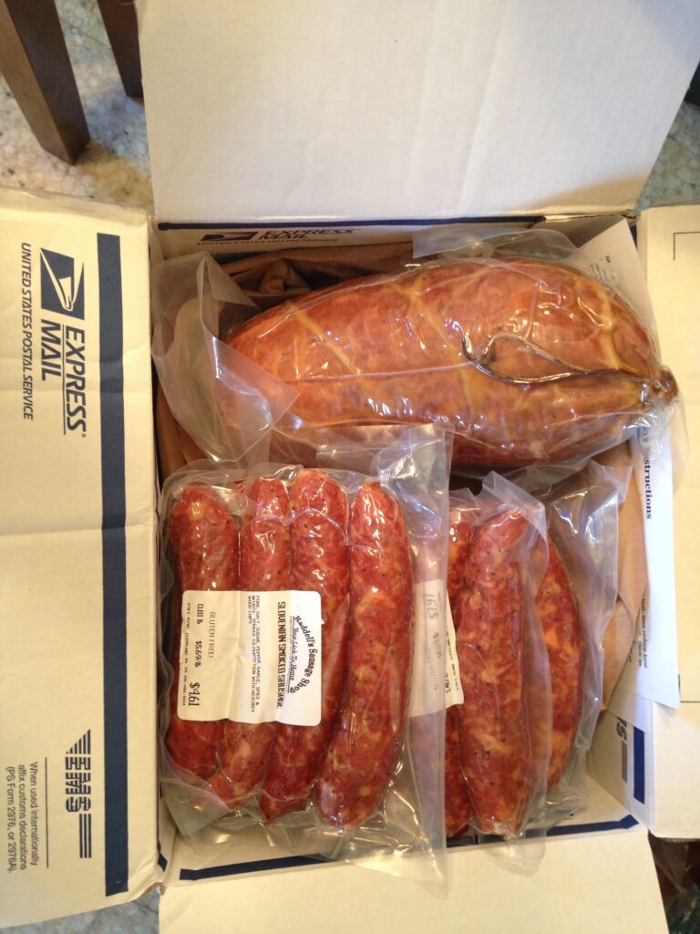 A cardboard box containing a large loaf of bread and several vacuum-sealed packages of cured, reddish sausages. The box is labeled "USPS Express Mail."
