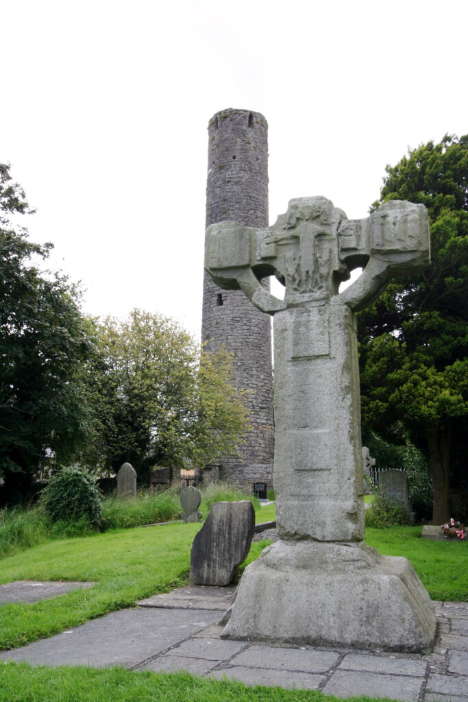 A large Celtic cross in a grassy cemetery with gravestones and an old stone tower in the background. The scene is serene, featuring green grass and trees under a cloudy sky.